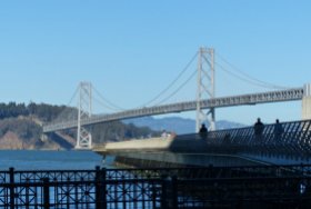 The bridge seen from the Embarcadero promenade. People strolling a pier—this one looks like a footbridge to the island.